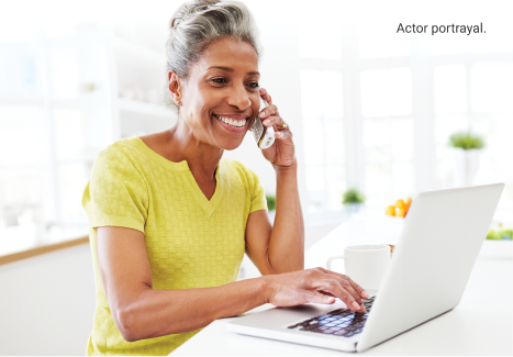 Actor portrayal of female patient in bright yellow blouse sitting at a table holding a phone to her ear while typing on a laptop